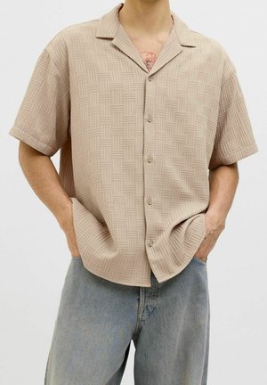 Osoba ubrana w beżową koszulę z krótkim rękawem z geometrycznym wzorem i jasnoniebieskie dżinsy, ręce w kieszeniach, neutralne tło.