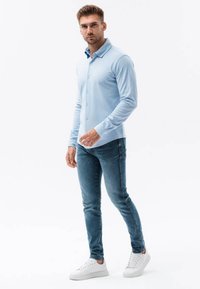 Camisa de manga larga azul claro con diseño de botones, combinada con unos jeans ajustados azules y zapatillas blancas. Tejido suave, corte ajustado.