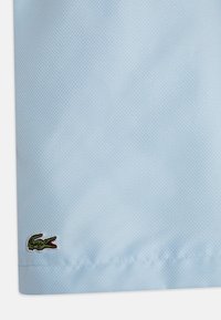 Camisa polo azul claro com um tecido texturizado, apresentando um pequeno logo de crocodilo verde bordado no canto inferior esquerdo.