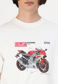 Hvid t-shirt med et grafisk print af en rød og grå motorcykel med etiketter, sat mod en ensfarvet baggrund.