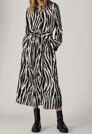 Woman wearing black and white zebra-striped long-sleeve belted dress with pockets, paired with black ankle boots on a neutral background.