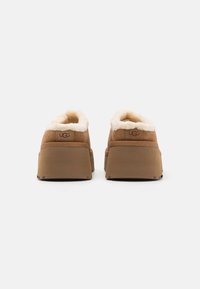 Zapatos de ante marrón tipo slip-on con un suave forro de color crema y suelas gruesas tipo plataforma. Presenta el logotipo UGG en relieve en el talón.