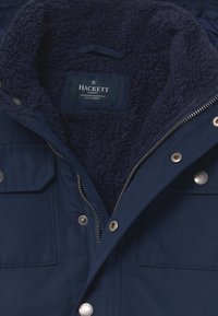 Giacca blu navy con fodera in pile morbido, chiusura con zip e bottoni a pressione, e due tasche frontali; etichetta del brand "HACKETT" visibile all'interno.