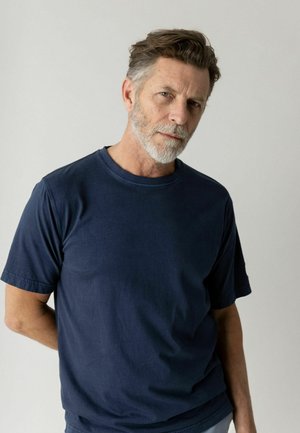 Keskeas vana mees hallide juuste ja habemega, kandes tumesinist T-särki, seisab käed selja taga ühtlase tausta ees.