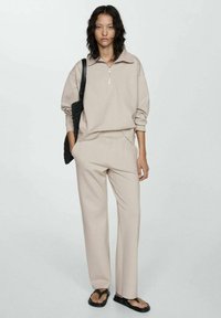 Pullover beige con zip e pantaloni abbinati in un materiale morbido e elasticizzato. Gli accessori includono una borsa tote nera intrecciata e sandali neri.