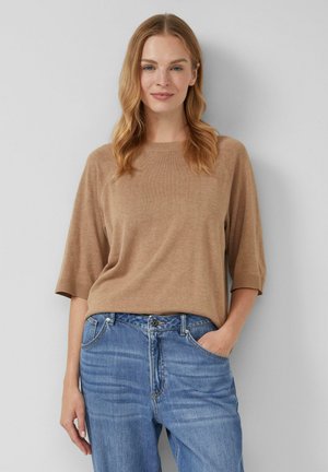 Kobieta z do ramion falującymi blond włosami, ubrana w beżowy sweter z krótkim rękawem i niebieskie dżinsy, stojąca z jedną ręką w kieszeni.