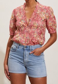Short-sleeved, pink floral blouse with ruffled neckline and buttons, paired with denim shorts featuring a rolled hem and pockets.
