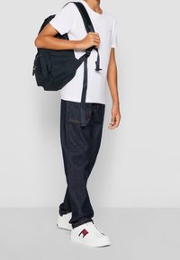 Mochila negra con múltiples compartimentos, camiseta blanca, pantalones vaqueros azul oscuro, zapatillas blancas con toques rojos y marinos. Conjunto casual y funcional.