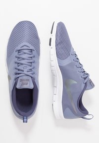 Lilla atletikskor i mesh med en tekstureret overdel, flade snørebånd og en hvid sål. Har et diskret metallisk logo på siden.