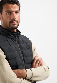 Schwarze gepolsterte Weste mit Reißverschluss vorne und kleinem Logo, getragen über einem strukturierten hellbeigen Pullover. Glatter Stoff mit leichtem Glanz.