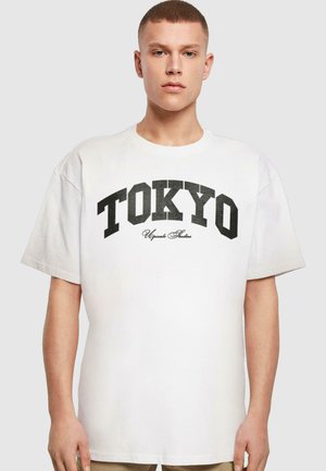 Fehér, laza szabású pamut póló, fekete "TOKYO" felirattal és alatta írott "Uganda Studios" logóval, kerek nyakkal és rövid ujjakkal.