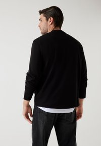 Maglia nera a maniche lunghe realizzata in tessuto morbido con scollatura rotonda, indossata sopra una camicia bianca, abbinata a jeans scuri. Design semplice, senza motivi.