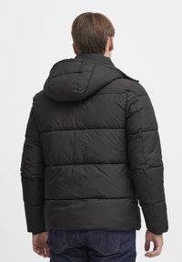 Sort pufferjakke med hætte, som har et quiltet design, vandrette sektioner og elastiske manchetter. Bæret med blå jeans.