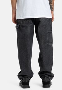 Czarne dżinsowe spodnie cargo z elastycznymi mankietami, z wieloma kieszeniami i lekko wyblakłą teksturą. Noszone z białą koszulką.