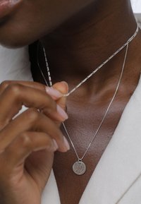 Colliers superposés en argent avec un pendentif circulaire texturé, tenu par une main. Le pendentif texturé et la fine chaîne ressortent bien sur le teint de la peau.