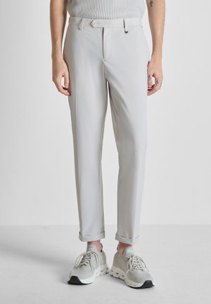 Persona che indossa pantaloni su misura grigio chiaro con cintura e sneakers grigio chiaro abbinate, in piedi davanti a uno sfondo semplice.