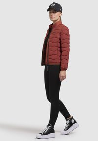 Rode pufferjack met golvend design, zwarte legging en hoge sneakers met witte zolen. Een zwarte pet met logo maakt de outfit compleet.
