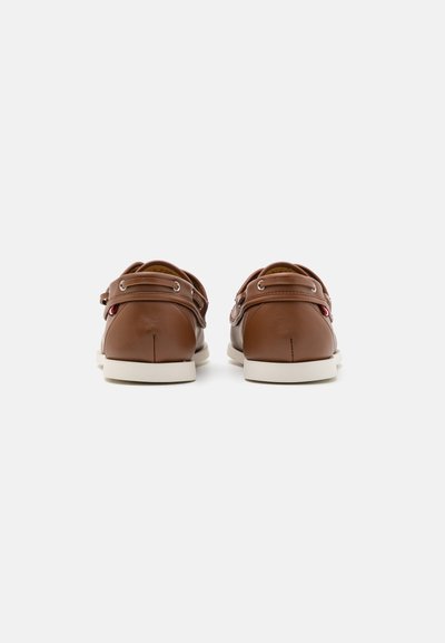 Pantofi din piele maro, cu vârful rotund, talpă din cauciuc alb și trei orificii cu accesorii metalice argintii de fiecare parte pentru închidere.