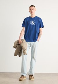 Camiseta de algodón azul con el logo blanco de "Calvin Klein", combinada con jeans de un azul claro y un sudadera beige sostenida en la mano. Zapatos de ante marrón.