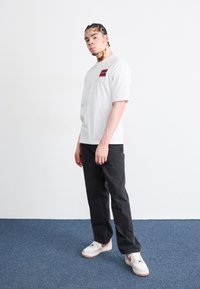 White t-shirt with a square red logo on the chest, black jeans, and white sneakers with pink accents. Plain background with blue floor.