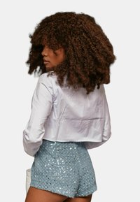 Camicetta bianca corta, pantaloni corti leggeri in blu con paillettes e dettagli argentati, che mostrano una superficie lucida e testurizzata. I capelli sono voluminosi e ricci.