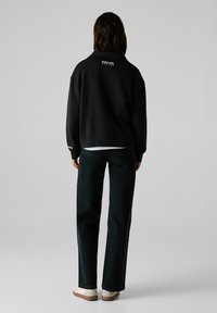 Sudadera negra de corte holgado, con un logo en la parte posterior, combinada con jeans de mezclilla oscura y zapatos de color crema.