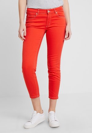 Femme portant un jean slim court rouge vif et des baskets blanches, debout sur un sol gris clair.