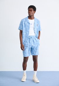 Lyseblå kortermet skjorte og matchende shorts med blomster mønster. Skjorten har en camp-krage; modellen har på seg en hvit T-skjorte under.