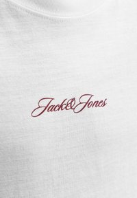 Λευκό ύφασμα με κόκκινο καλλιγράφημα λογότυπο "Jack & Jones" τυπωμένο κοντά στον γιακά σε ένα μπλουζάκι.