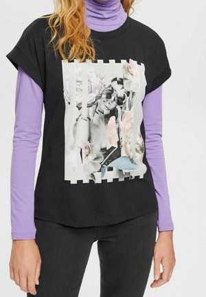 Sort t-shirt med et sort-hvidt foto af en kvinde med solbriller, omgivet af blomster i pastelfarver, båret over en lyserød/lilla langærmet trøje.