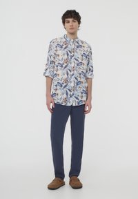 Camicetta a maniche corte con colletto che presenta un motivo floreale blu e beige su sfondo bianco, abbinata a pantaloni blu scuro e scarpe slip-on marroni.
