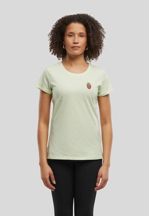 Femme aux cheveux bouclés portant un t-shirt vert pâle avec un petit motif de fraise sur la poitrine et un pantalon noir, debout devant un fond uni.
