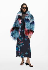 Flauschiger, mehrfarbiger Kunstpelzmantel in Blau-, Rosa- und Lilatönen; kombiniert mit einem dunkelblumigen Kleid und schwarzen, hochhakigen Sandalen.