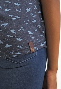 Mörkblå t-shirt med ljusblå fågelmönster, korta ärmar, rund halslinning och en brun etikett vid nederkanten. Slätt tyg, avslappnad passform.