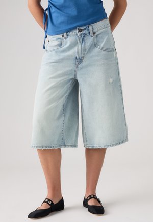 Persona con pantalones cortos vaqueros de pierna ancha hasta la rodilla de color azul claro, top azul y zapatos planos negros con tiras decoradas con ojales.