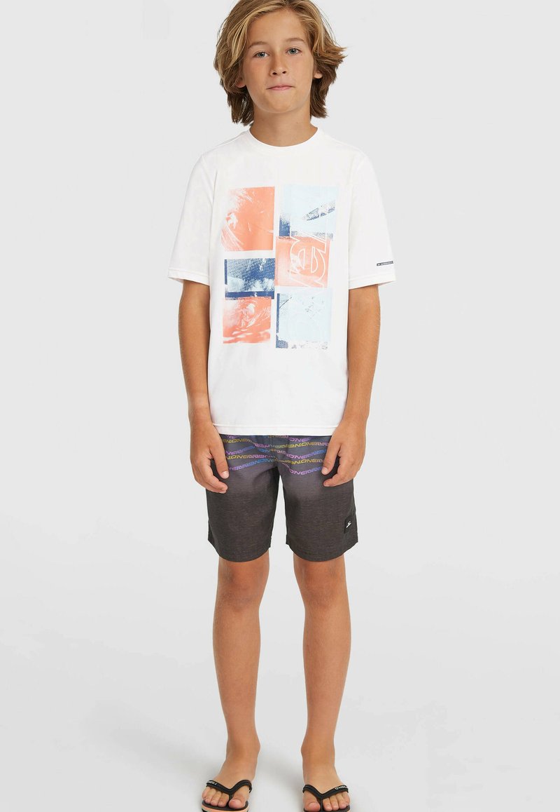 Mladý chlapec stojí, má na sebe biele tričko s potlačou, čierne šortky s farebnými pruhmi a čierne šľapky, na jednoduchom pozadí.