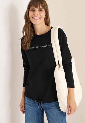 Zwarte langemouwen top met de tekst "PURE REFLECTION" in grijs. Gecombineerd met lichtblauwe jeans en een crèmewitte gewatteerde schoudertas.