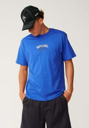 Joven con una camiseta azul de Rip Curl, gorra negra con logo blanco, pantalones negros, mirando hacia abajo con las manos en los bolsillos.