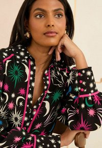 Svart blus med flerfärgade stjärn- och månmönster, med rosa detaljer och knapputskrifter. Långa ärmar med åtsittande manschetter.