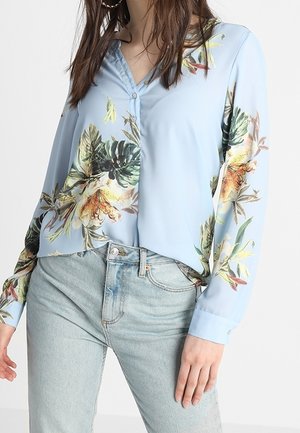 Blusa azzurra chiara con un motivo floreale tropicale, dotata di un'apertura con bottoni davanti, maniche lunghe e una vestibilità comoda. Abbinata a jeans in denim chiaro.