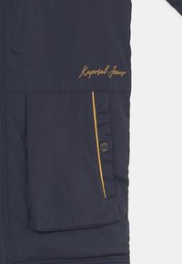 Námořnická modrá bunda s hladkým povrchem, s zlatou výšivkou "Kaporal Jeans", žlutým akcentním lemem a kapsou na cvok.