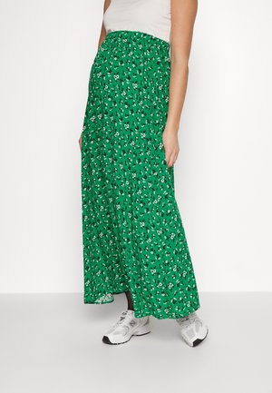 Jupe maxi verte avec un motif floral, dotée d'une taille élastique ajustée et d'un tissu fluide jusqu'aux chevilles, associée à des baskets blanches.