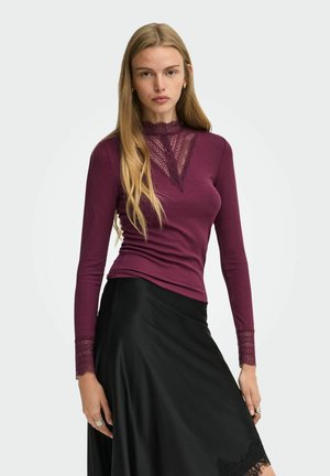 Jeune femme aux longs cheveux blonds portant un haut violet à manches longues avec bordure en dentelle et une jupe noire sur un fond uni.