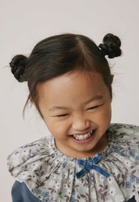 Dítě oblečené v blůze s květinovým vzorem, s volánovým límečkem a modrou sametovou mašlí, široce se usmívající. Vlasy jsou upravené do dvou drdolů.