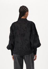 Blusa nera trasparente con maniche a sbuffo e motivi floreali ricamati. Presenta polsini con bottoni e dettagli texturizzati. Indossata sopra jeans grigi.