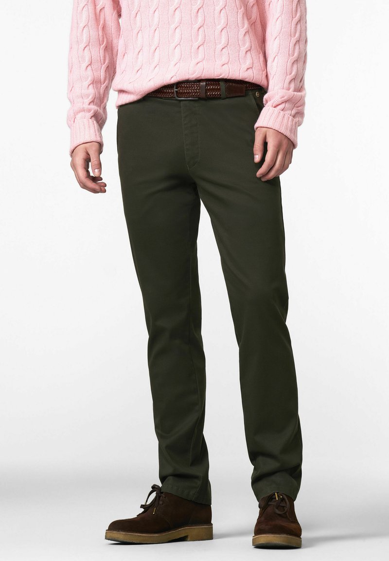 Homme portant un pantalon vert foncé, une ceinture marron, des chaussures en daim marron et un pull en tricot torsadé rose clair, se tenant devant un fond blanc.