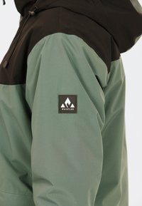 Grüne und schwarze Jacke mit Kapuze, die eine strukturierte Oberfläche hat. Sie verfügt über ein Logo-Patch am Ärmel mit einem Bergdesign.