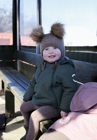 Egy barna bojtos sapkát és zöld cipzáras dzsekit viselő gyerek fa padon ül, közelében barna leggings és egy rózsaszín dzseki.