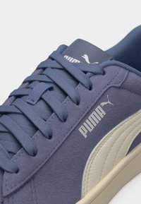 Κοντινό πλάνο μοβ αθλητικού παπουτσιού Puma με κορδόνια στο ίδιο χρώμα, λευκό λογότυπο Puma και ρίγα, που δείχνει υφή από σουέντ και δερματικά υλικά.