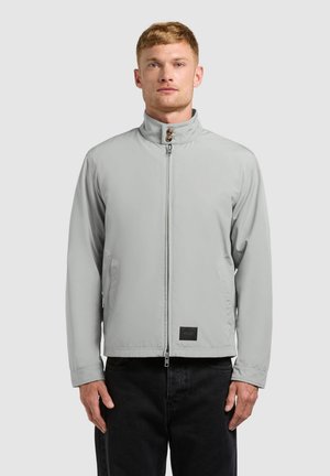 Mann trägt eine hellgraue Jacke mit Reißverschluss und hochgeschlagenem, mit Knöpfen versehenem Kragen sowie schwarze Hose, steht vor einem schlichten hellgrauen Hintergrund.